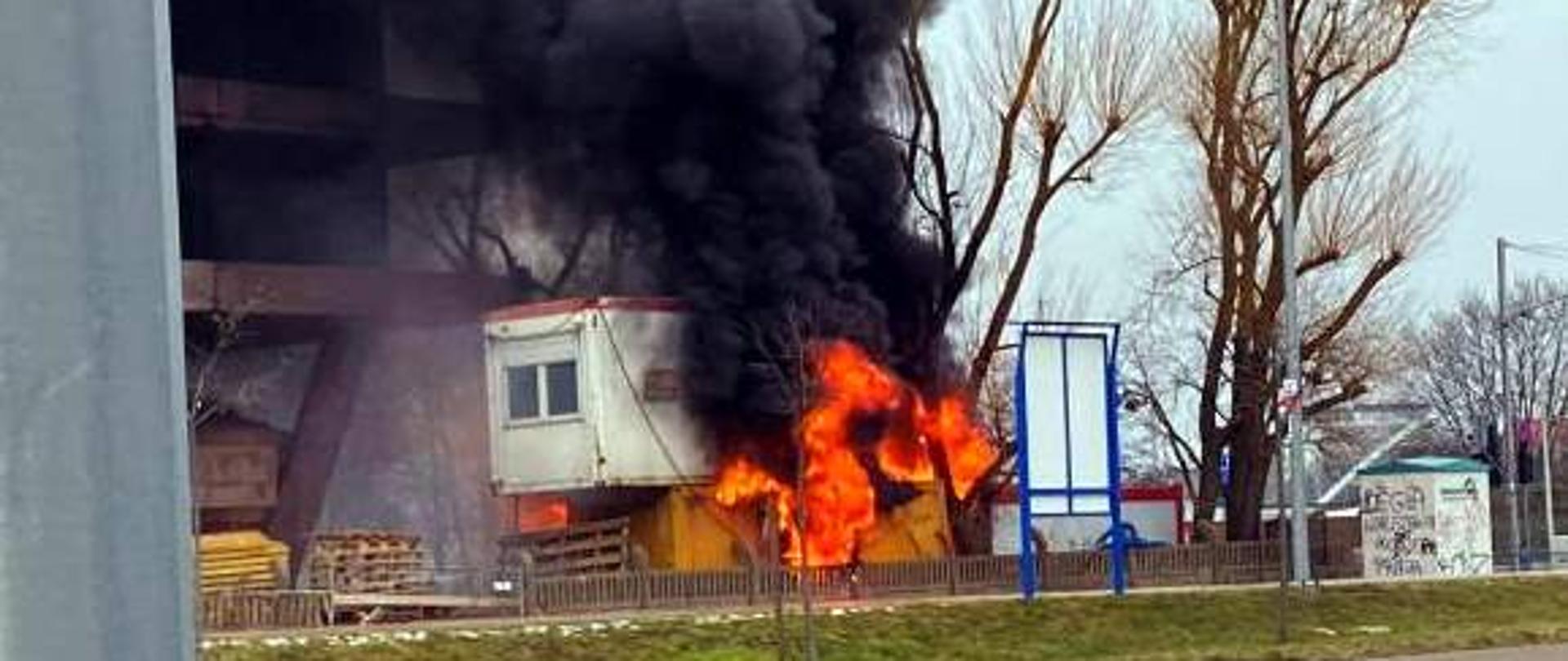 Kontenery objęte ogniem, unoszący się dym, po lewej nowobudowany obiekt mieszkalny, na placu budowy składowane drewniane palety. W oddali drzewa.