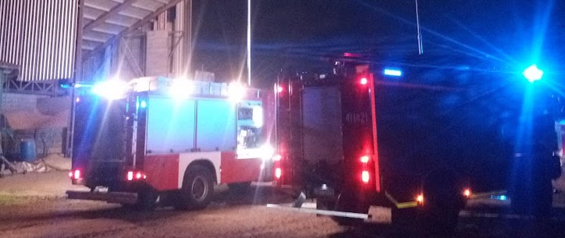 Zdjęcie przedstawia samochody ratowniczo-gaśnicze z włączonymi niebieskimi światłami ostrzegawczymi oraz wysuniętymi masztami oświetleniowymi podczas akcji gaszenia pożaru suszarni do zboża 