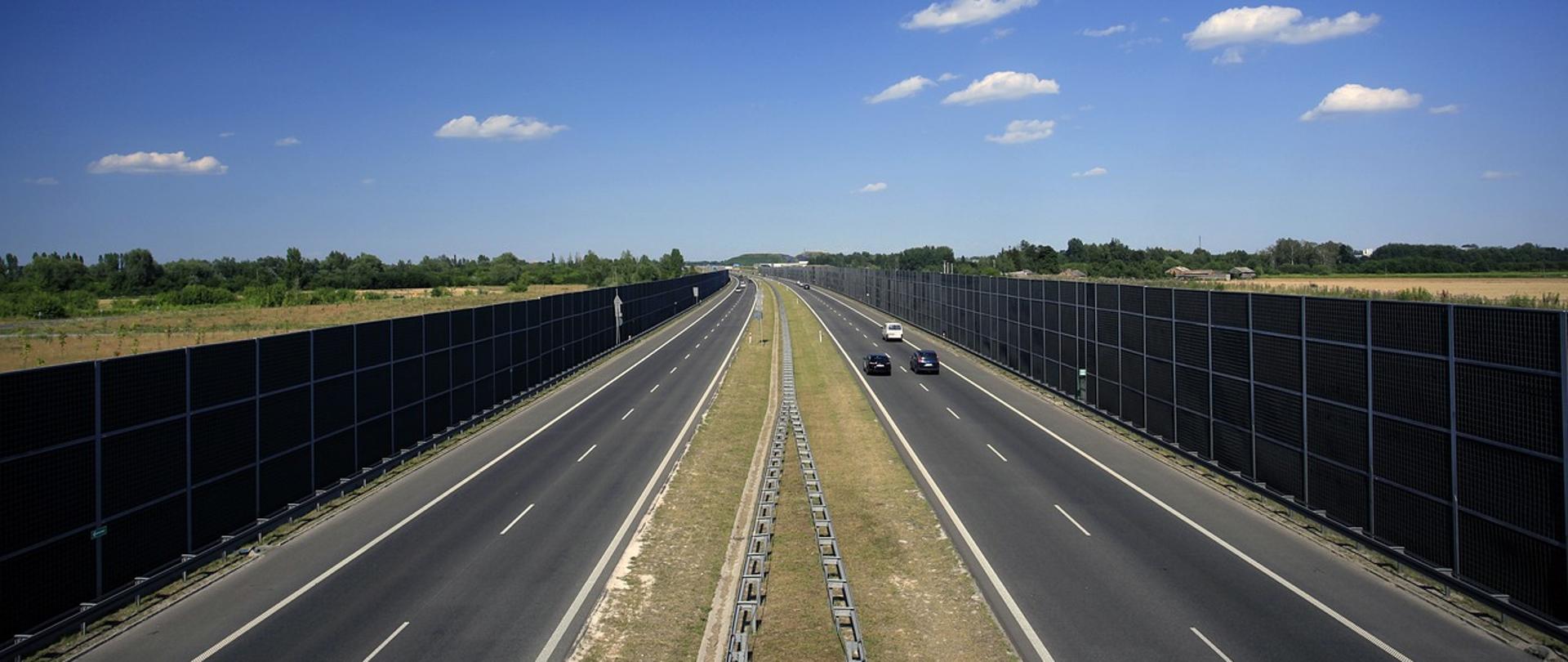 Autostrada po której jadą samochody. Po obu stronach autostrady są ekrany akustyczne. 