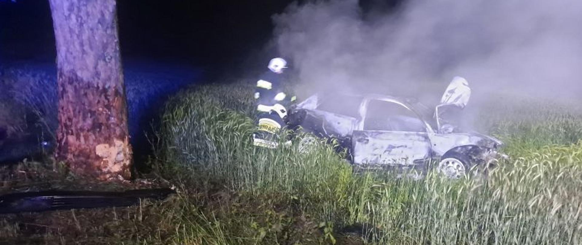 Zdjęcie nocne. Po lewej stronie zdjęcia drzewo. Drzewo w miejscu uderzenia auta bez kory. Po prawej auto. Auto dymi się, jest koloru srebrnego. Przy aucie strażacy w ciemnych mundurach i białych hełmach.