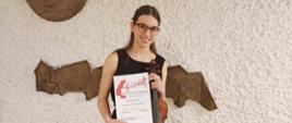 Zdjęcie kolorowe. Uczennica w czarnej sukience trzyma w rękach skrzypce i dyplom Ogólnopolskiego Konkursu "Talenty Muzyczne 2023". Na białej ścianie za nią zawieszone dwie płaskorzeźby z mosiądzu - po lewej stronie okrągła płaskorzeźba z wizerunkiem Mieczysława Karłowicza, poniżej płaskorzeźba przedstawiająca pasmo górskie.