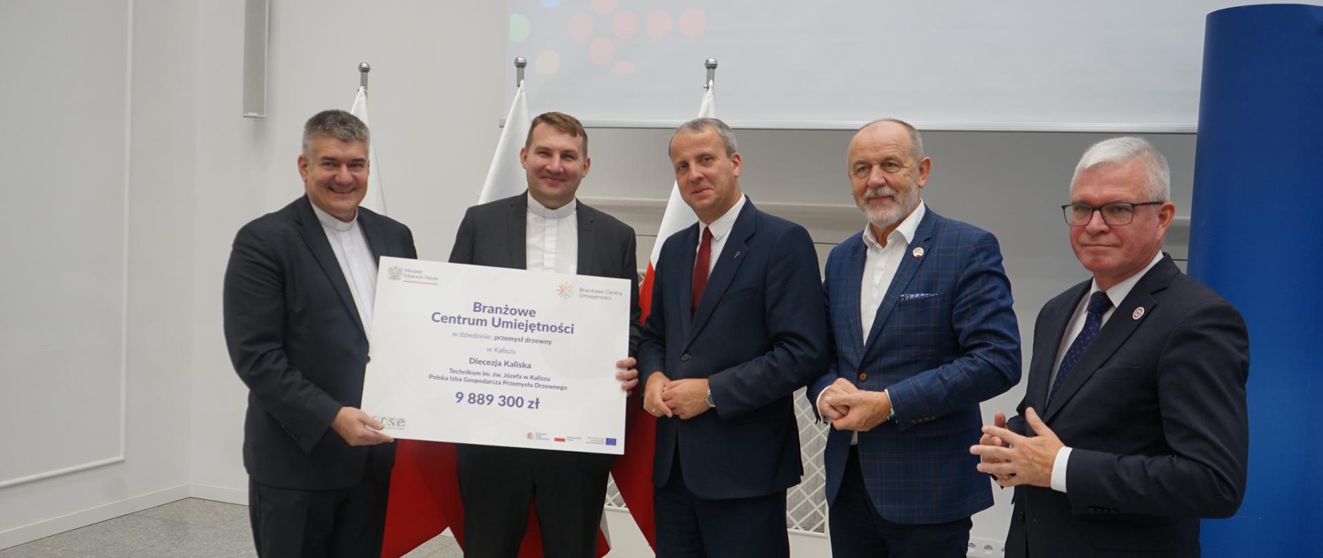 Pod białą ścianą stoi pięciu mężczyzn w garniturach, dwóch z lewej trzyma wielki symboliczny czek z napisem Branżowe Centrum Umiejętności - 9 889 300 zł.