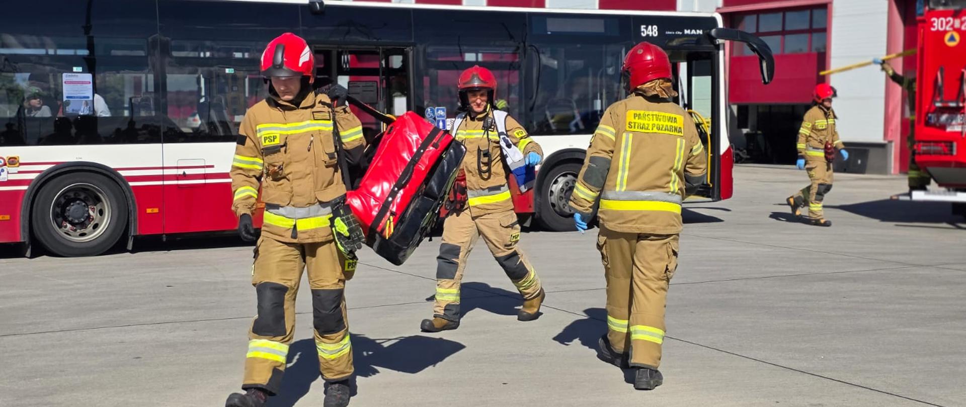 Kilku strażaków PSP idzie wzdłuż autobusu i wozu strażackiego, niosąc torby medyczne i sprzęt ratowniczy. W tle widoczne są budynki zajezdni. Scena pokazuje przygotowanie sił i środków do działań w ramach zdarzenia masowego.