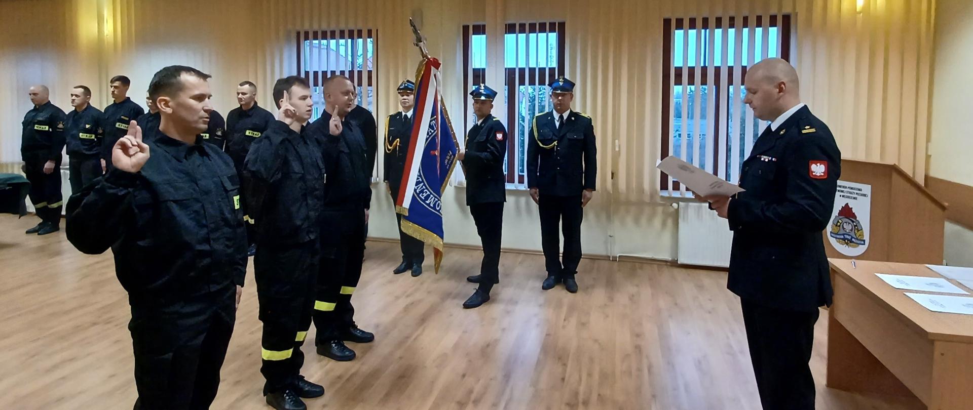 Na sali narad odbywa odbywa się uroczyste ślubowanie 3 strażaków na sztandar. Za nimi widać innych strażaków stojących w rzędzie 