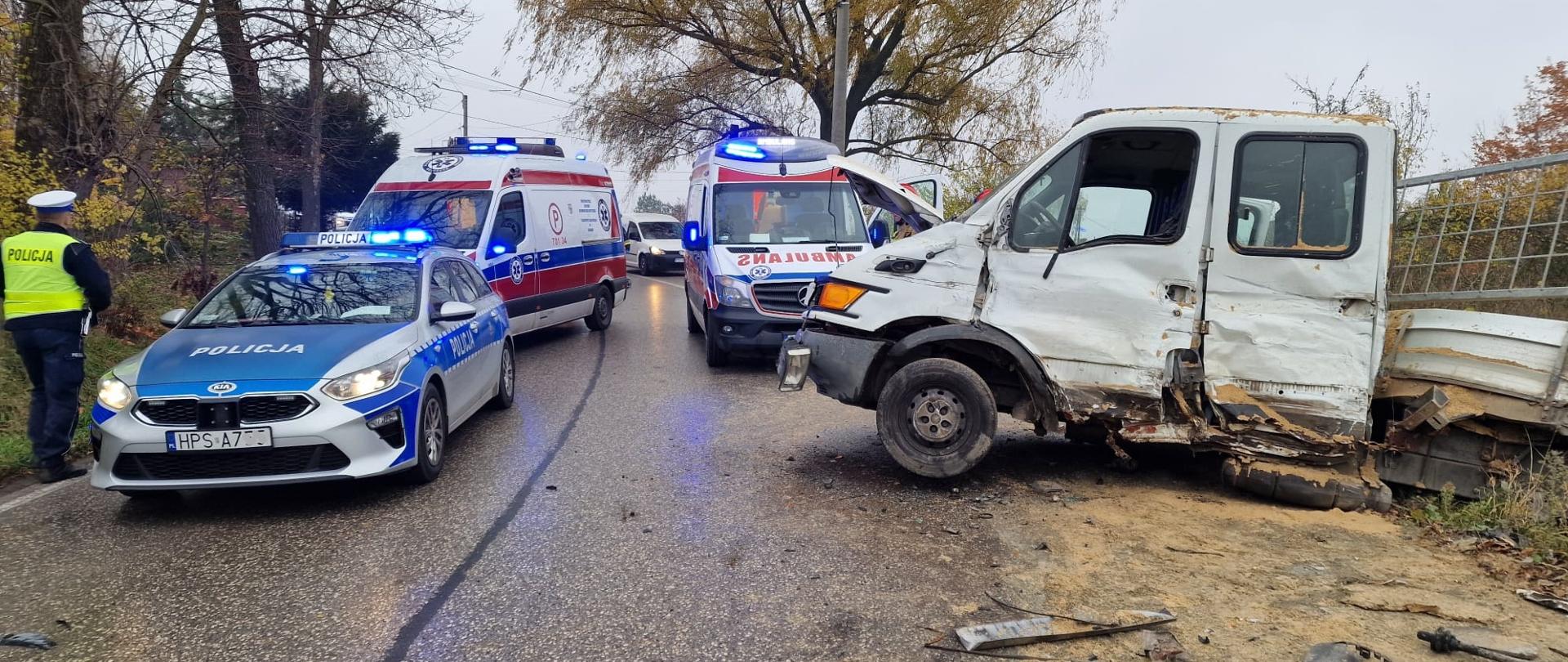 Rozbity bok samochodu dostawczego częściowo na jezdni, w tle policjant i pojazdy służb ratowniczych