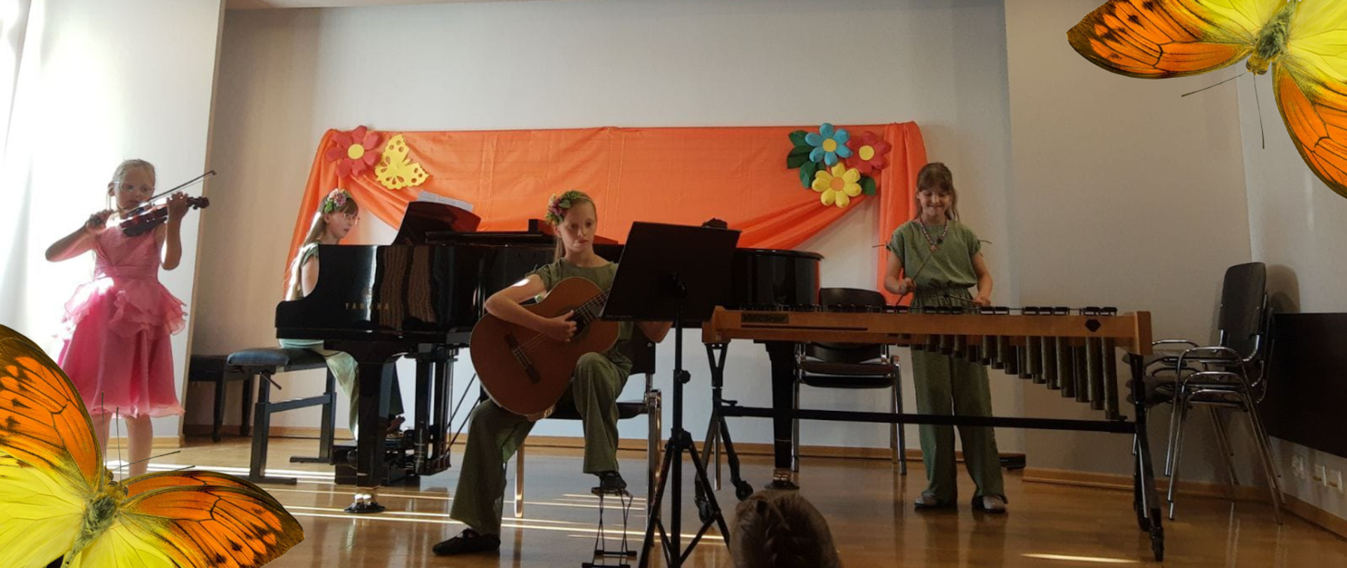 Na scenie czworo uczniów. Same dziewczęta. Jedna z nich gra na gitarze, inna na perkusji, kolejna na fortepianie. Te trzy dziewczęta są ubrane w bluzki i spodnie w kolorze zielonym. Ostatnia dziewczyna gra na skrzypach. Jest blondynką ubraną w różową balową sukienkę. Sceneria zdjęcia bardzo wiosenna w tle motyli i kolorowe kwiaty.