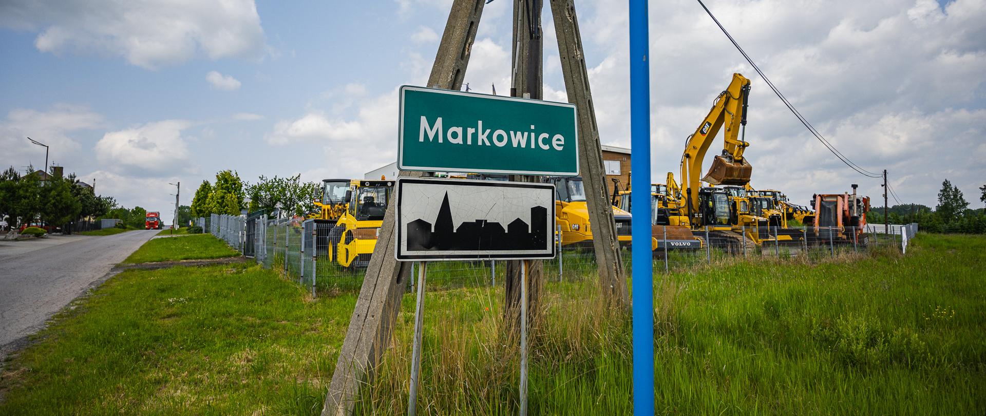Znak miejscowości „Markowice” ustawiony przy lokalnej drodze. Nad nim znajduje się tablica z nazwą ulicy „ul. Św. Floriana”. W tle widoczny jest ogrodzony plac z maszynami budowlanymi, w tym koparkami i walcami drogowymi, oraz częściowo zachmurzone niebo.