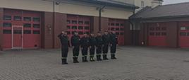 Strażacy stoją na placu podczas zbiórki z okazji odzyskania przez Polskę niepodległości. Za nimi w tle i po prawej stronie budynek z czerwonymi wrotami garażowymi.