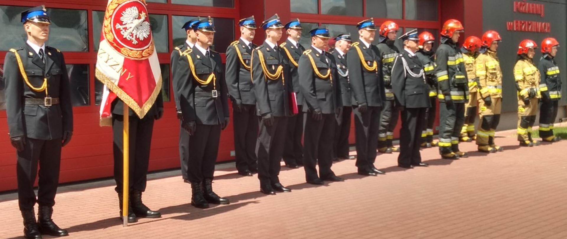 na zdjęciu plac przed budynkiem Komendy Powiatowej PSP w Brzezinach, stoją pododdziały strażaków komendy na czele poczet sztandarowy komendy podczas uroczystej zbiórki z okazji Dnia Strażaka