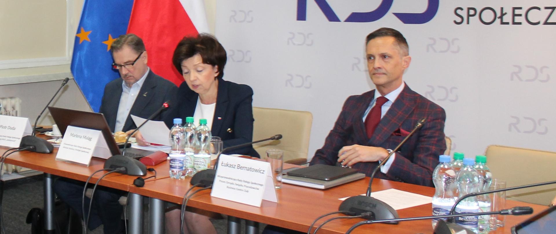 Na zdjęciu widnieje stół prezydialny za którym siedzą od lewej strony: Piotr duda – przewodniczący NSZZ Solidarność, Marlena Maląg – Minister Rodziny i Polityki Społecznej, Przewodnicząca Rady Dialogu Społecznego, Łukasz Bernatowicz prezes zarządu BCC. W tle siedzących znajduje się baner z napisem Rada Dialogu Społecznego oraz flagi.