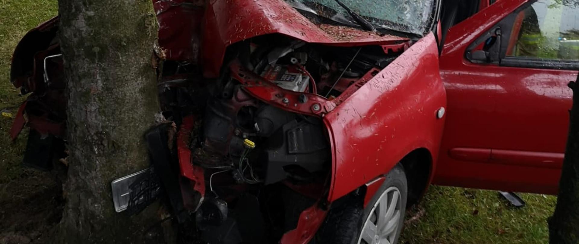 Samochód koloru czerwonego przodem uderzył w drzewo. W tle budynek mieszklany