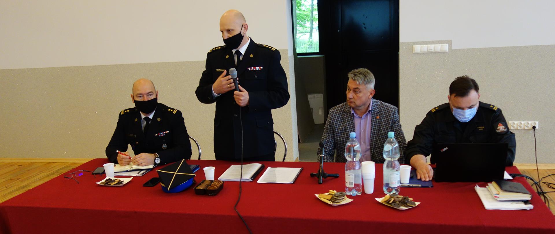 Przy stole nakrytym czerwonym obrusem siedzą Komendanci PSP w Kutnie z przedstawicielami OSP - w trakcie konsultacji. Stojąc przemawia trzymając mikrofon Komendant Powiatowy Państwowej Straży Pożarnej w mundurze wyjściowym.