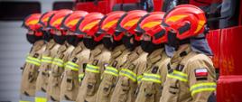 Na zdjęciu widoczni strażacy zmiany służbowej w umundurowaniu piaskowym specjalnym w czerwonych hełmach za nimi czerwone samochody pożarnicze podczas Powiatowych obchodów Dnia Strażaka w KP PSP w Gorlicach. 