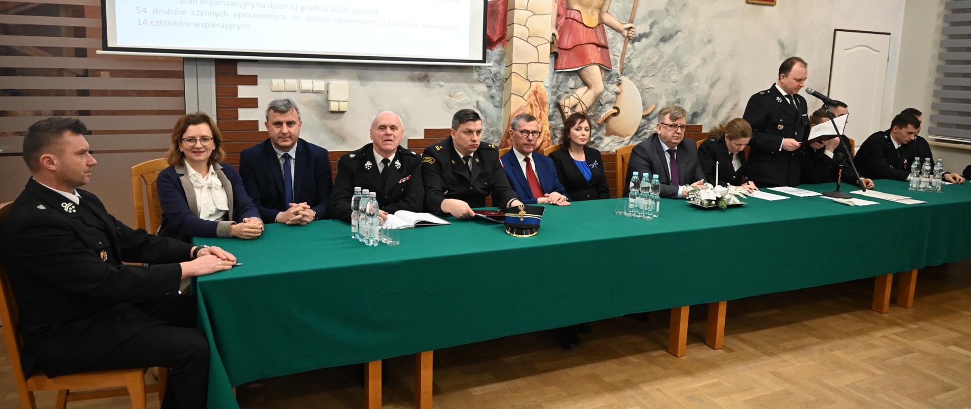 Zdjęcie przedstawia stół przy którym siedzą zaproszeni goście oraz organizatorzy spotkania między innymi dowódca Jednostki Ratowniczo-Gaśniczej Numer 1 w Kielcach mł. bryg. Damian Domański. W tle płaskorzeźba Świętego Floriana.