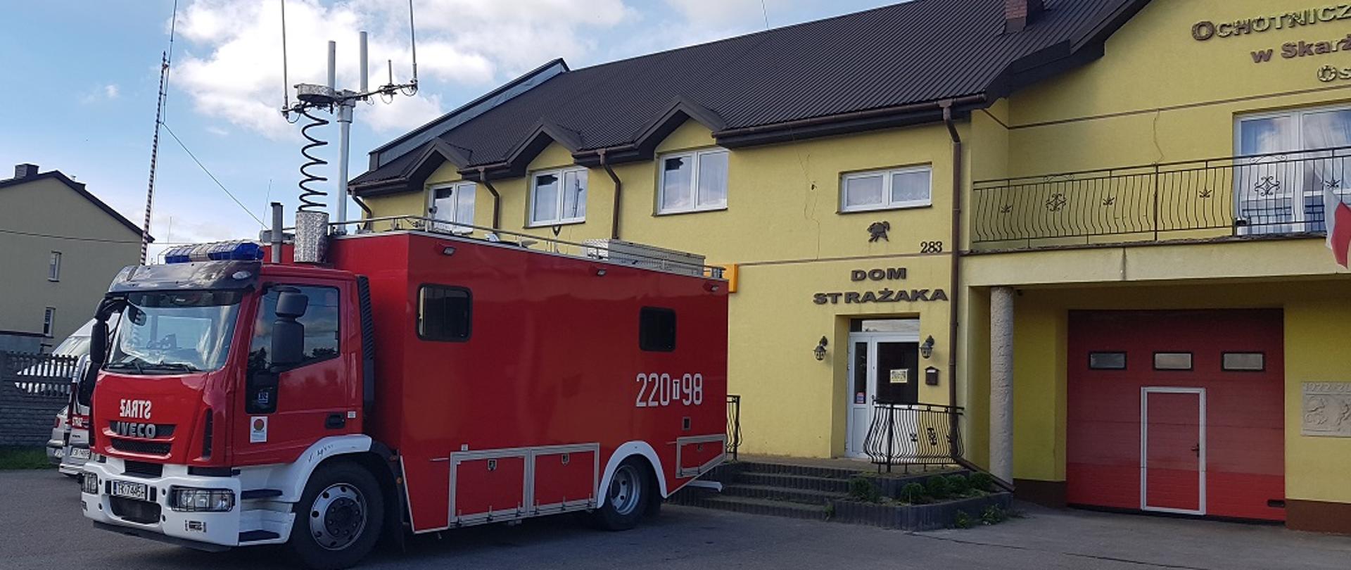 Na zdjęci widać czerwony pojazd państwowej Straży Pożarnej na tle żółtego budynku. Widoczny pojazd jest samochodem dowodzenia i łączności widoczne są na nim wysunięte anteny. Pojazd stoi na podjeździe wykonanym z kostki brukowej. W tle widać budynek remizy z brązowym poszyciem dachem wykonanym z blachy. Elewacja budynku jest w kolorze żółtym. 
