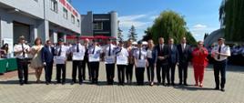 Przedstawiciele jednostek ochotniczych straży pożarnych i zaproszeni goście stoją na placu manewrowym przed strażnicą. W tle budynek i samochód strażacki oraz pozostali uczestnicy uroczystości.