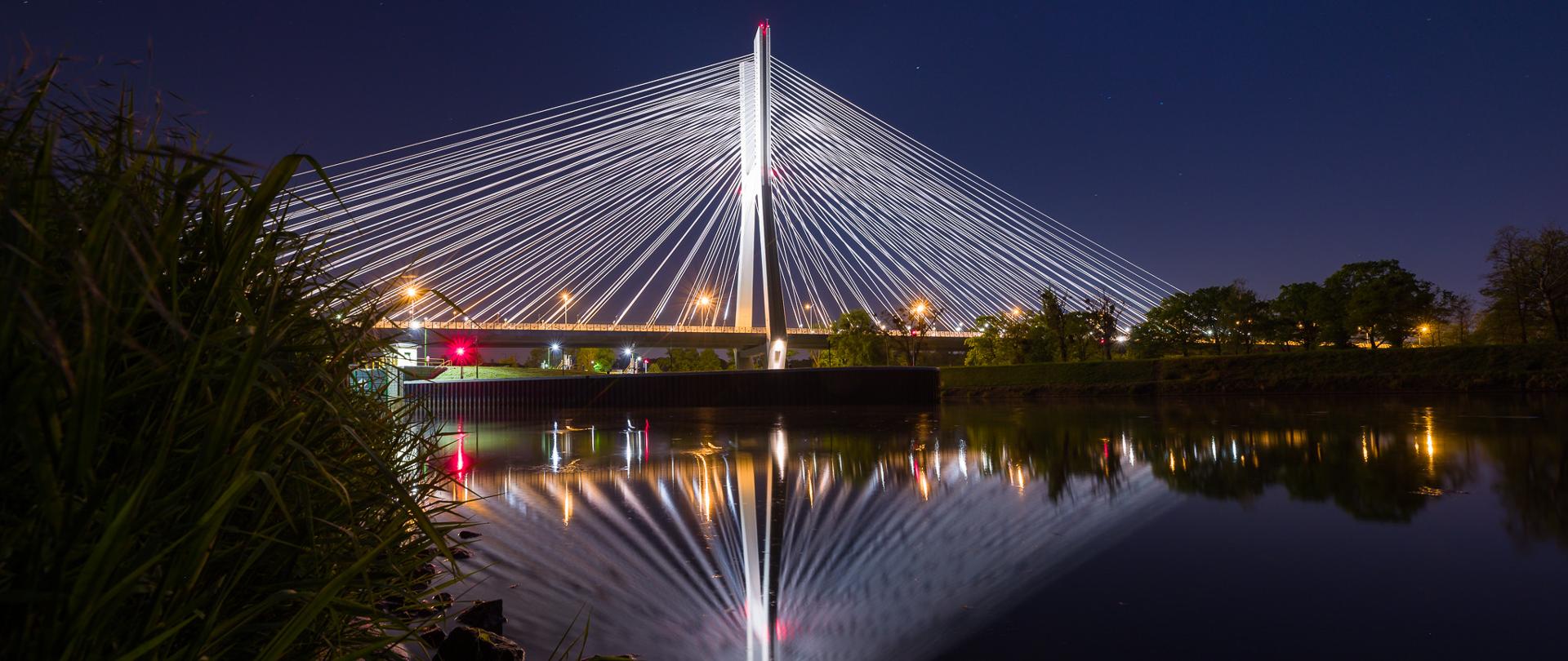 Podświetlony Most Rędziński w nocy. Zdjęcie z poziomu rzeki. Na rzece malowniczo odbijają się elementy mostu.