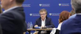 Wiceminister Jacek Karnowski siedzi w środku przy stole i mówi do mikrofonu.