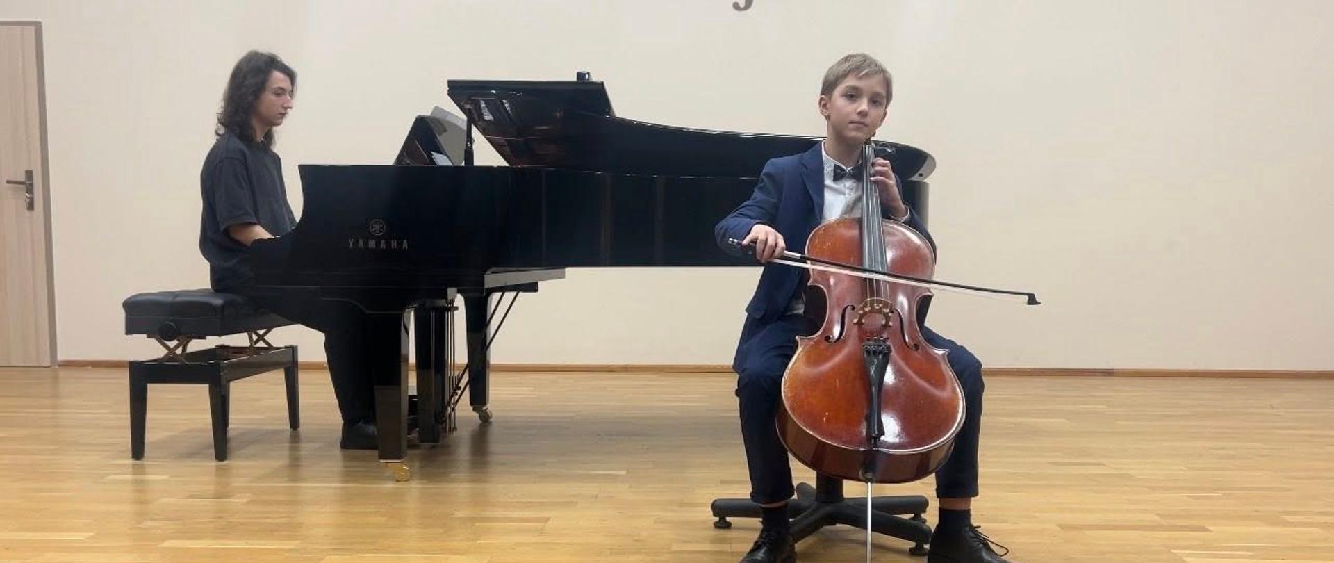 Na sali koncertowej gra wiolonczelista. W tle przy fortepianie akompaniator.