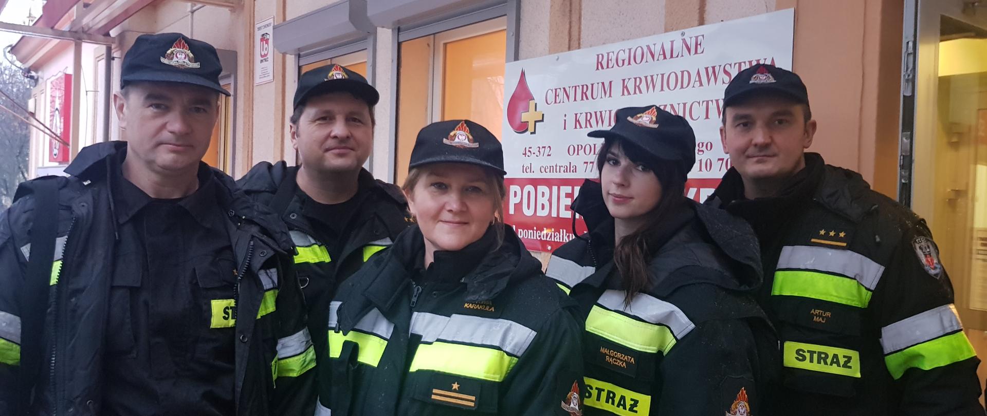 Strażacy PSP przed budynkiem RCKiK w Opolu, tuż przed oddaniem krwi. Strażacy w ubraniach dowódczo-sztabowych i koszarowych z czarnymi czapeczkami na głowach.