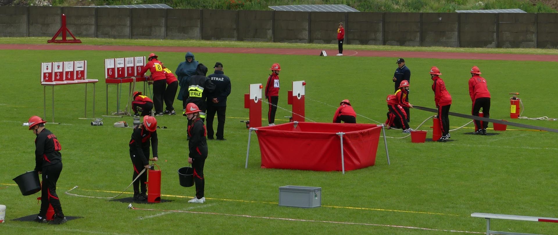 Zdjęcie przedstawia przebieg Zawodów Młodzieżowych Drużyn Pożarniczych Powiatu Brodnickiego wg regulaminu CTIF. Dzieci rywalizują na murawie stadionu w konkurencji rozwinięcie bojowe.