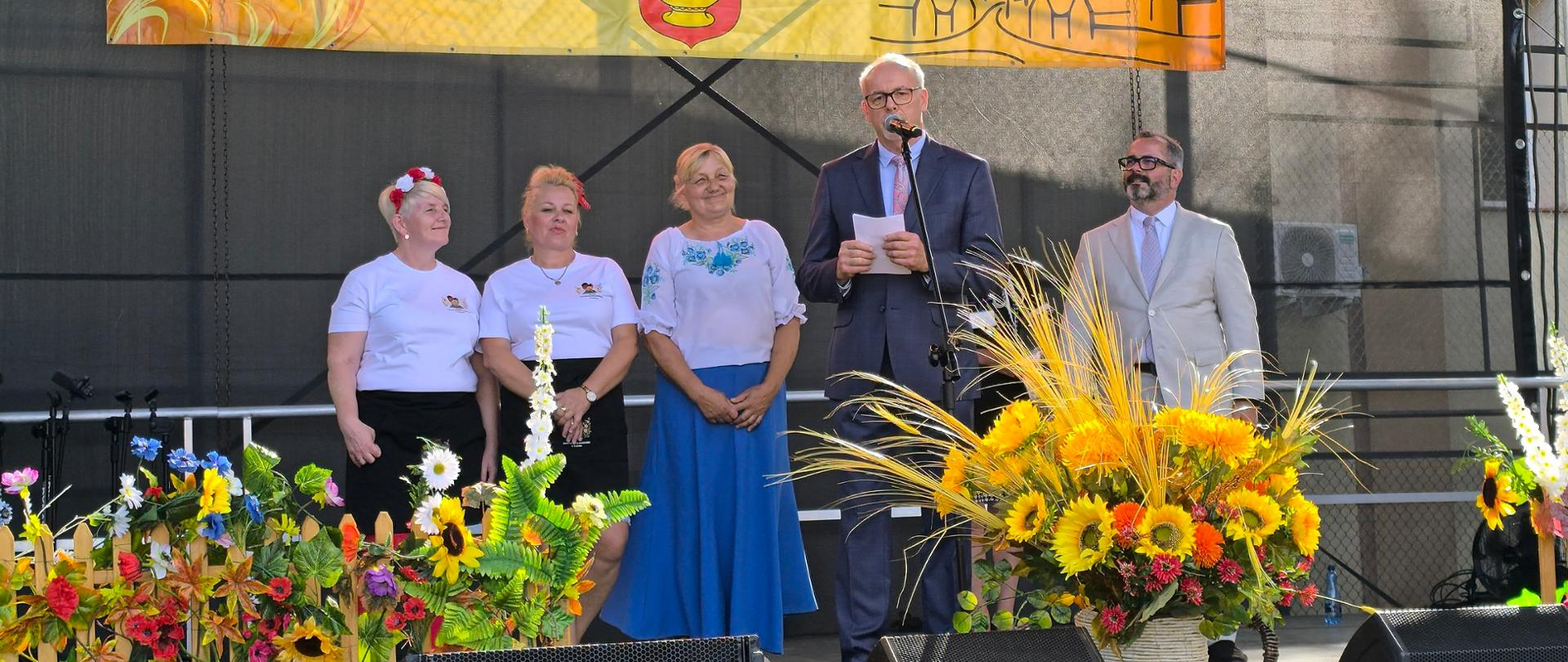 Trzy kobiety i dwaj mężczyźni stoją na scenie udekorowanej jesiennymi kwiatami, w tle plakat Dożynki Gminne Bielsk.