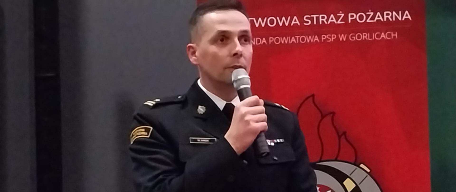 Zajęcie przedstawia przemawiającego z mikrofonem komendanta powiatowego Państwowej Straży Pożarnej w Gorlicach podczas narady rocznej podsumowującej działalność Komendy Powiatowej Państwowej Straży Pożarnej w Gorlicach.