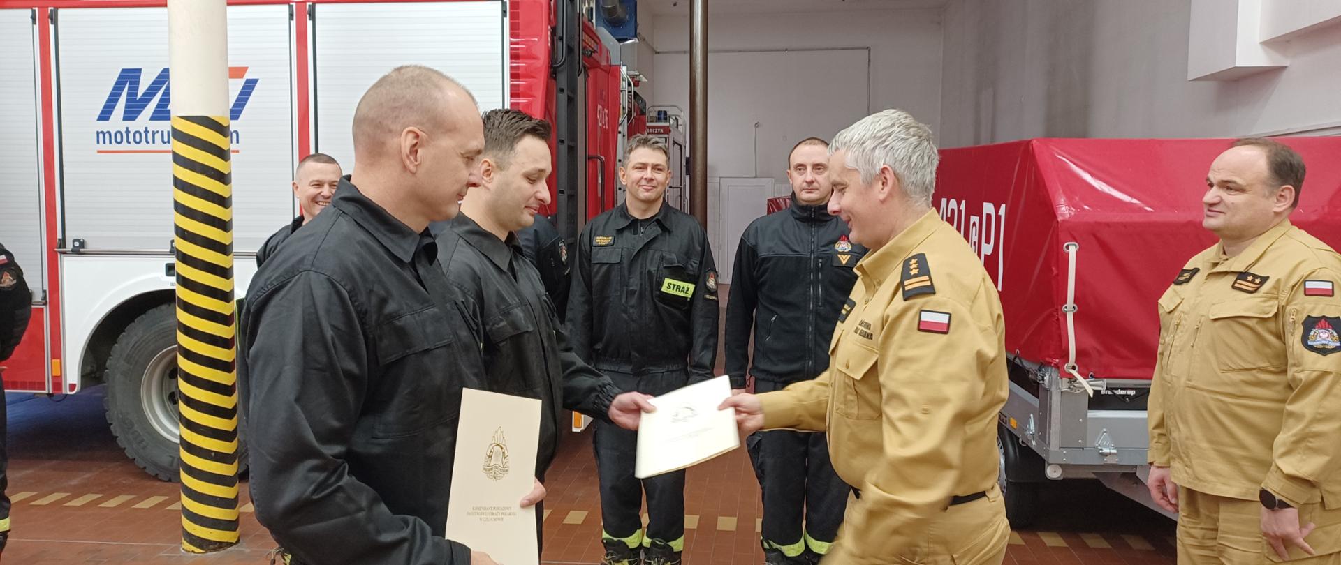 Zdjęcie przedstawia Komendanta Powiatowego Państwowej Straży Pożarnej w Człuchowie st. bryg. Radosława Batruch wręczającego nagrodę finansową ogn. Damianowi Chojnackiemu i st. str. Dawidowi Synoradzkiemu. Za nimi Zastępca Komendanta Powiatowego PSP w Człuchowie bryg. Henryk Koźlewicz i strażacy stojący w rzędzie w pomieszczeniach garażowych jednostki, w tle samochody pożarnicze.