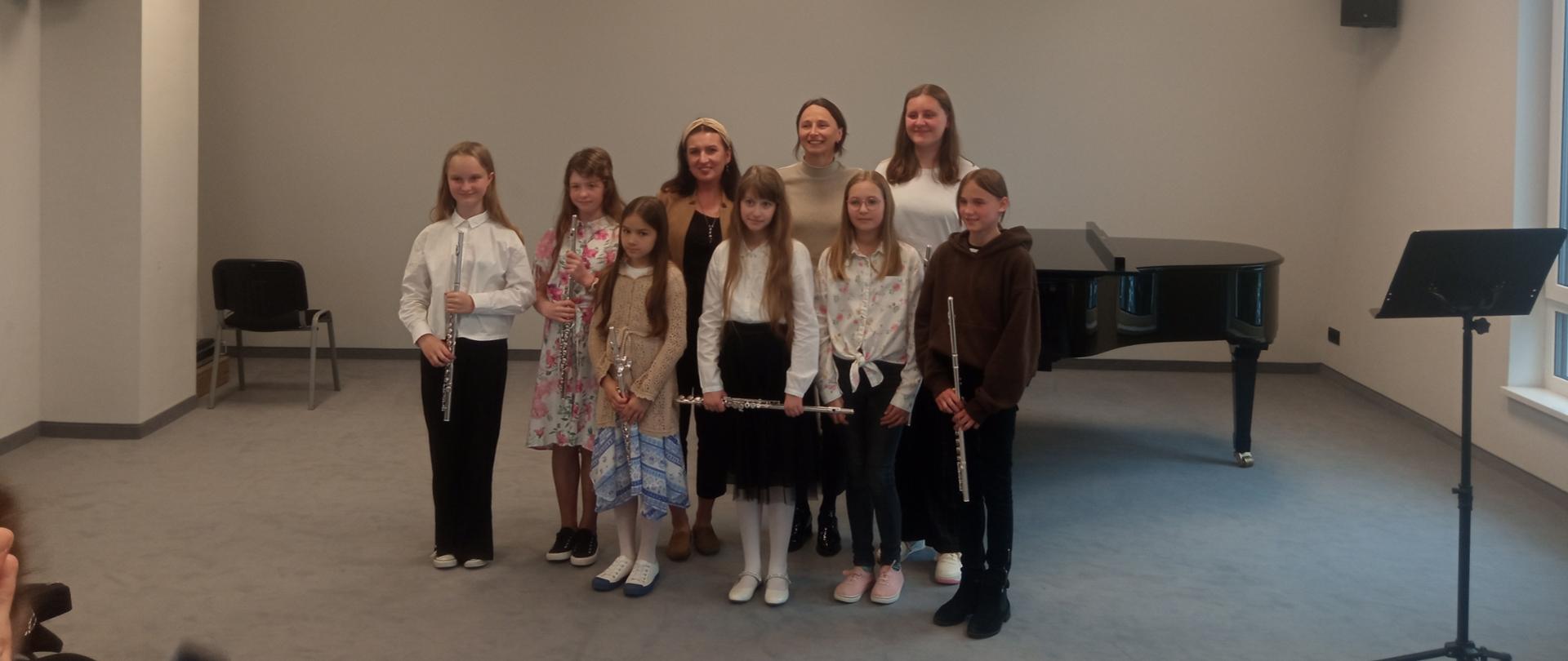 Klasa fletu pani Magdaleny Margańskiej-Dec , dziewczynki stojące wraz z panią nauczycielką i akompaniatorką trzymają w dłoniach flety, po prawej stronie pulpit, z tyłu po lewej stronie krzesło. Za pozującymi do zdjęcia fortepian. 