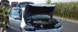 Kolizja w Żylicach. Na jezdni, na tle pola kukurydzy i budynków stoi auto osobowe. Za nim znajduje się Karetka pogotowia ratunkowego, obok widoczni są dwaj strażacy.