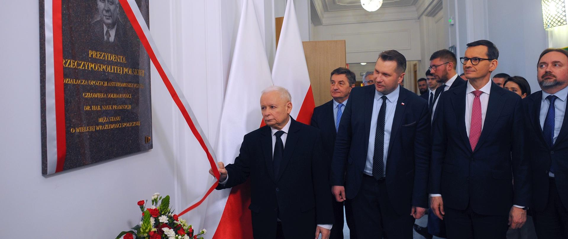 Widok na korytarz, na białej ścianie wisi ciemnoszara tablica z portretem Lecha Kaczyńskiego przesłonięta biało-czerwoną wstążką, przed nią stoi grupa mężczyzn w garniturach.