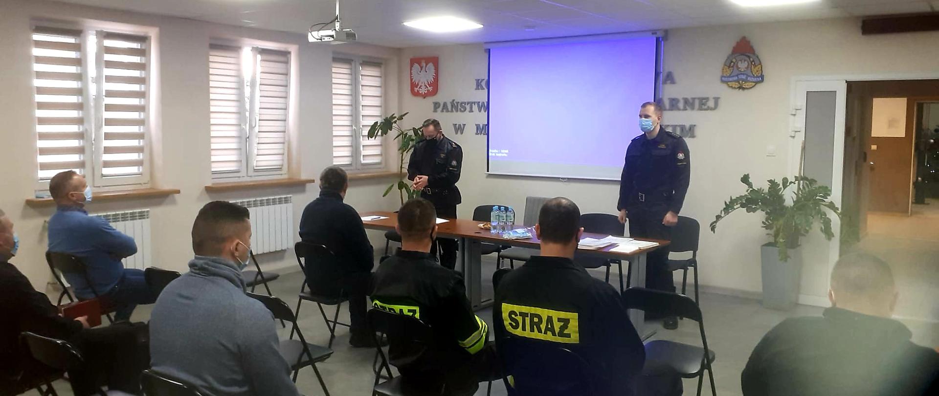Strażacy OSP siedzą na świetlicy na przeciwko stołu, za którym stoi dwóch funkcjonariuszy PSP. Na ścianie jest rozwinięty ekran od rzutnika oraz godło Polski i logo PSP. 