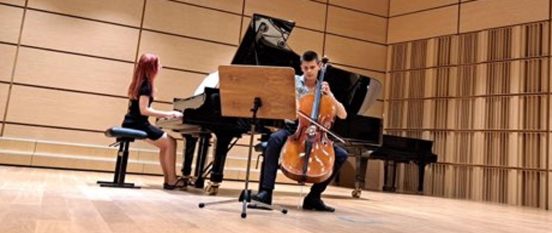Pianistka grająca na fortepianie wraz z towarzyszeniem wiolonczelisty na sali koncertowej. Przed wiolonczelistą widnieje pulpit.