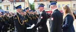 Na pierwszym planie oficer w ciemnym mundurze galowym przekazuje dokument strażakowi stojącemu w rzędzie na baczność. Obok nich stoi kobieta w cywilnym stroju wyjściowym oraz kolejny oficer, asystujący przy ceremonii. W tle widać długi szereg umundurowanych strażaków oraz zabytkową zabudowę miejską.