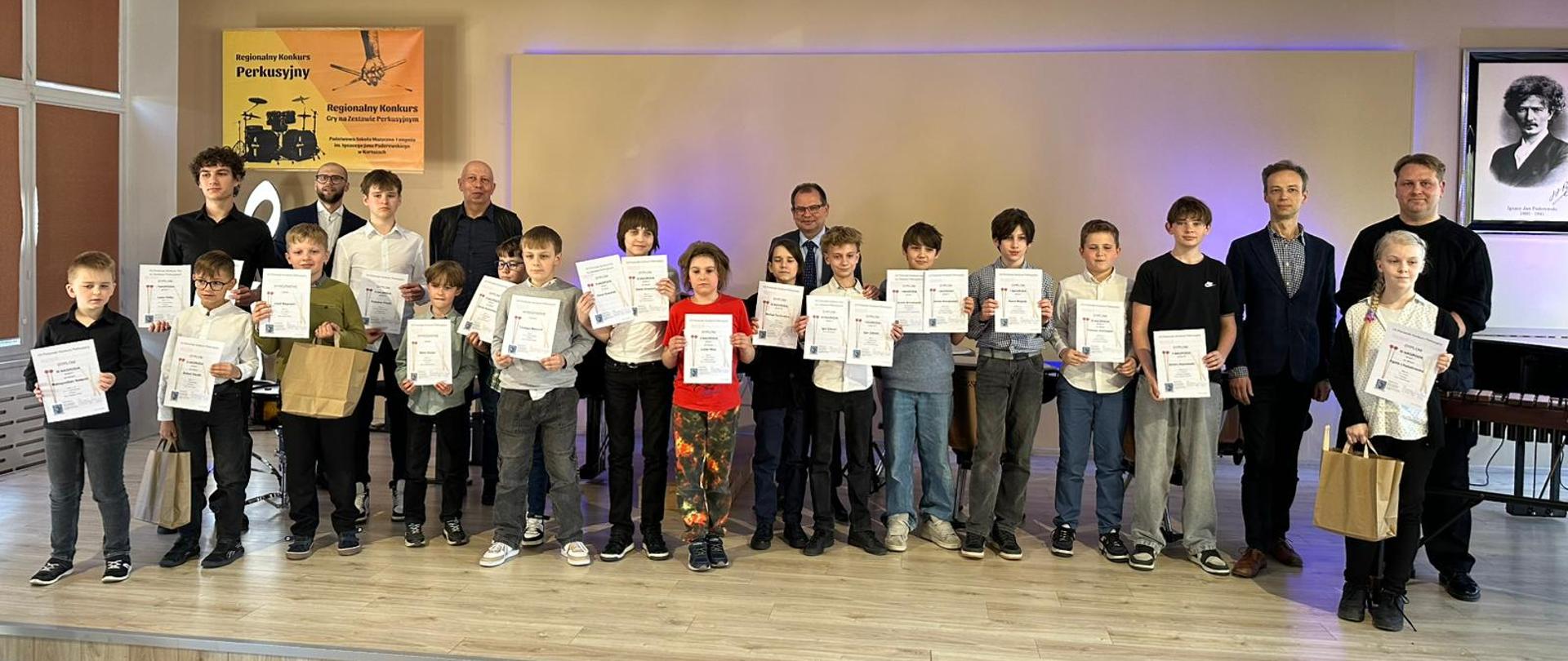 Na scenie duża grupa uczniów, którzy zostali laureatami konkursu perkusyjnego. Wszyscy trzymają dyplomy. Tło obrazka w odcieniach beżu.