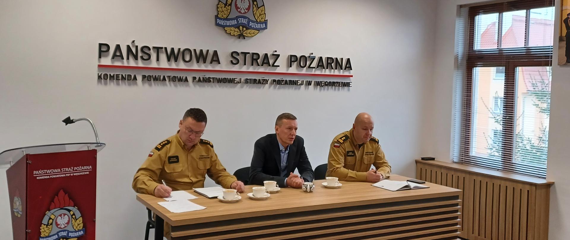 Zastępca Komendanta Wojewódzkiego PSP w Olsztynie wraz z przedstawicielem NFOŚ z Olsztyna oraz Komendant Powiatowy PSP w Węgorzewie siedzą przy biurku na sali konferencyjnej. 