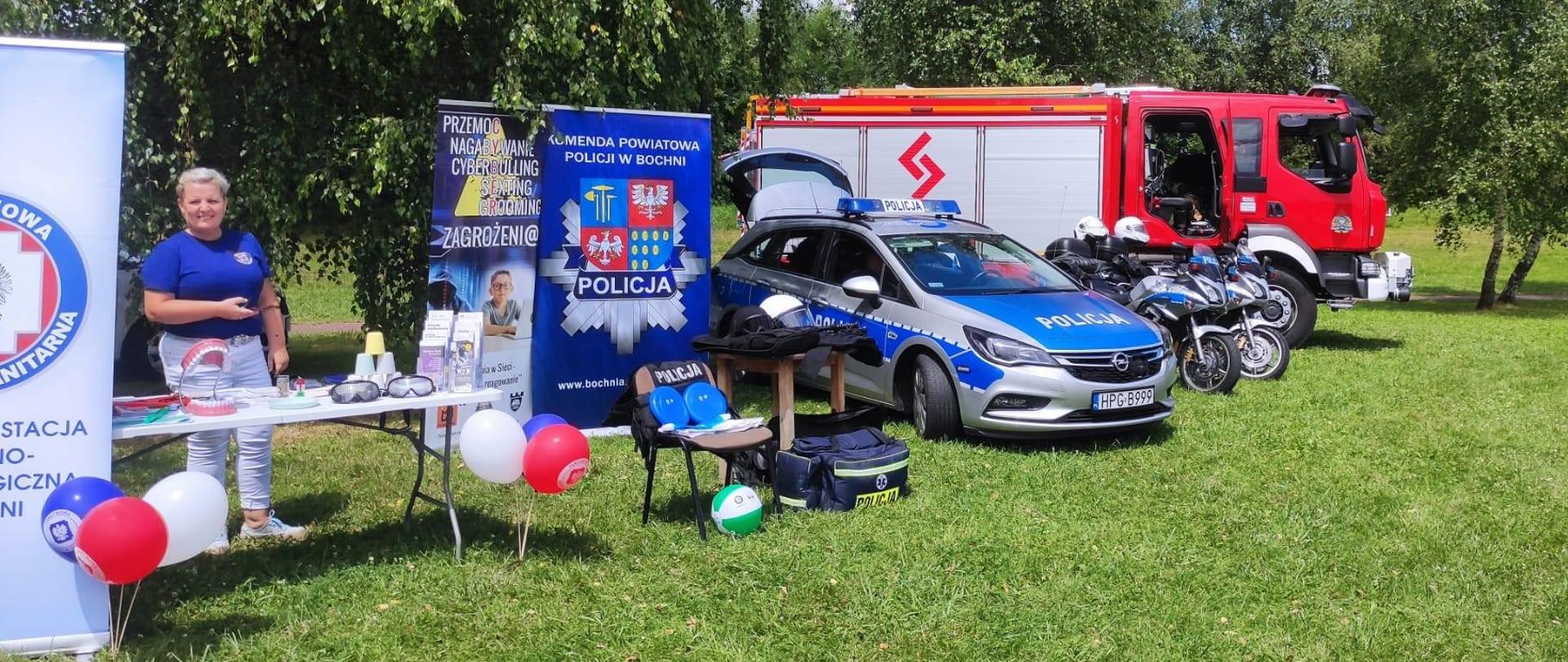 Zdjęcie przedstawia służby i inspekcję biorące udział w wydarzeniu. Na tle trawy i drzew znajduje się samochód strażacki, motocykle policyjne oraz samochód policyjny. Na pierwszym planie widać banery służb i inspekcji oraz stanowisko przygotowane przez Państwową Stację Sanitarno-Epidemiologiczną