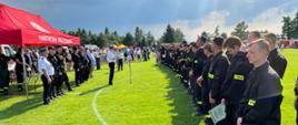 Zdjęcie przedstawia druhów OSP ustawionych z prawej strony w dwuszeregu. Ubrani są w ubranie koszarowe i czapki strażackie. Niektórzy z nich w dłoniach trzymają dyplomy i puchary. Przed strażakami stoi czerwony namiot z napisem Państwowa Straż Pożarna, a przed nim stoją władze miasta Biała Rawska oraz komisja sędziowska i Zastępcę Komendanta Wojewódzkiego PSP, który przemawia przez mikrofon. Dookoła stoją osoby cywilne. Wszystko dzieje się na boisku do piłki nożnej.