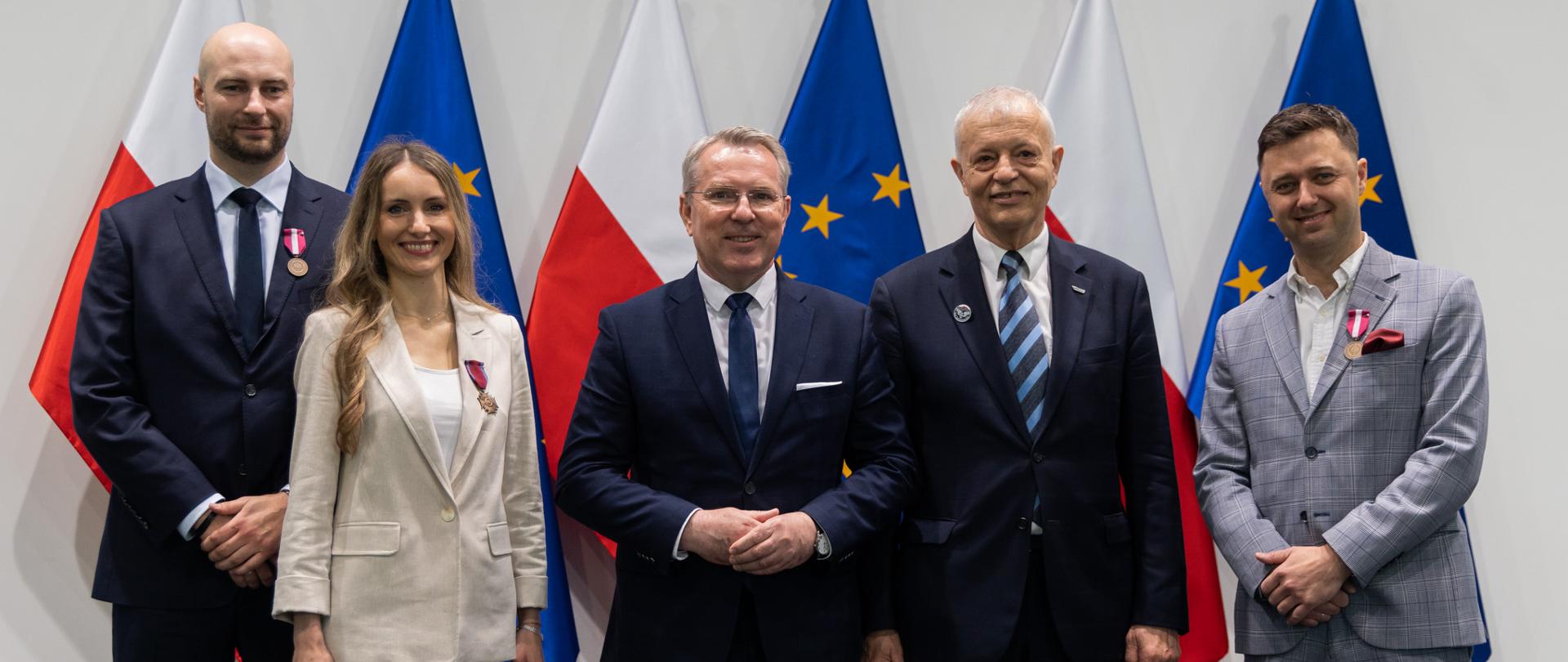 Zdjęcie grupowe z Wicewojewodą i innymi odznaczonymi. Na zdjęciu widoczne są flagi polski i unii europejskiej. 