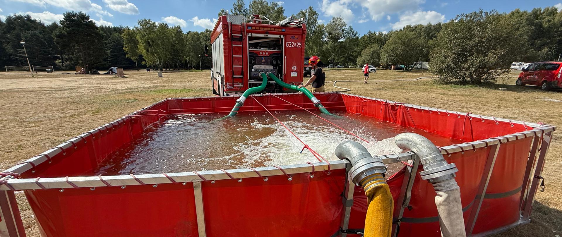 Na zdjęciu widać duży, przenośny basen retencyjny w kolorze czerwonym, wypełniony wodą. Do basenu pompowana jest woda za pomocą grubych węży pożarniczych, które są podłączone do wozu strażackiego widocznego w tle. Wokół basenu i w oddali znajdują się strażacy w mundurach, którzy nadzorują akcję. Całość rozgrywa się na otwartym, trawiastym terenie, w słoneczny dzień z chmurami na niebie. Basen retencyjny tego typu jest wykorzystywany przez straż pożarną jako tymczasowy zbiornik wody podczas akcji, szczególnie tam, gdzie nie ma łatwego dostępu do hydrantów.