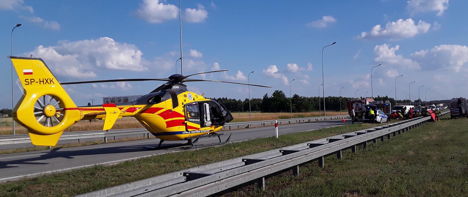 Zdjęcie przedstawia ratowników straży pożarnej oraz Zespołu Ratownictwa Medycznego podczas udzielania pomocy poszkodowanej osobie w wypadku na autostradzie A1. Z lewej widoczne są samochody straży pożarnej, z prawej karetka pogotowia. Na pierwszym planie widoczny jest śmigłowiec Lotniczego Pogotowia Ratunkowego. 