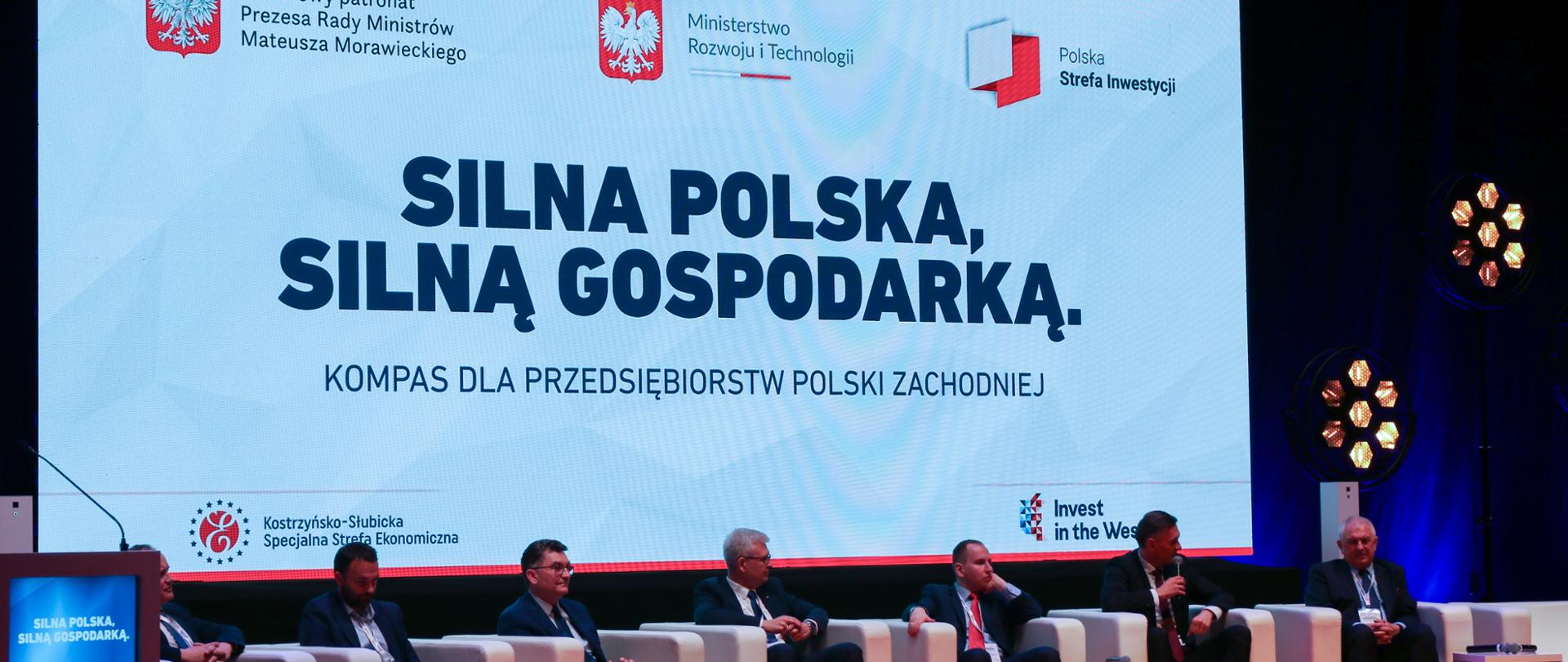 Uczestnicy konferencji siedzą na scenie w fotelach. Jeden obok drugiego i mówią do publiczności.