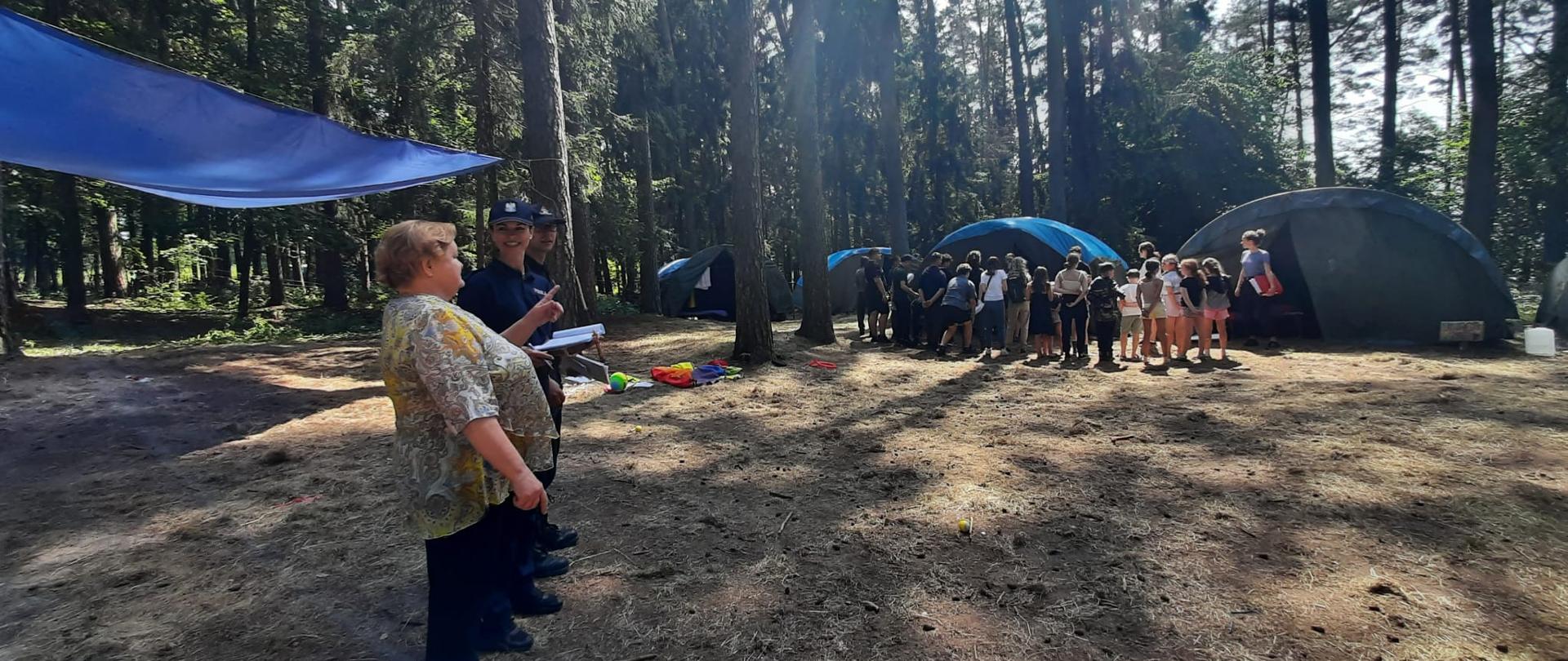 Strażak z policjantkom i starostą powiatu węgorzewskiego stoi w obozowisku. I przygląda się ewakuacji harcerzy. Harcerze stoją na zbiórce przed namiotem. 