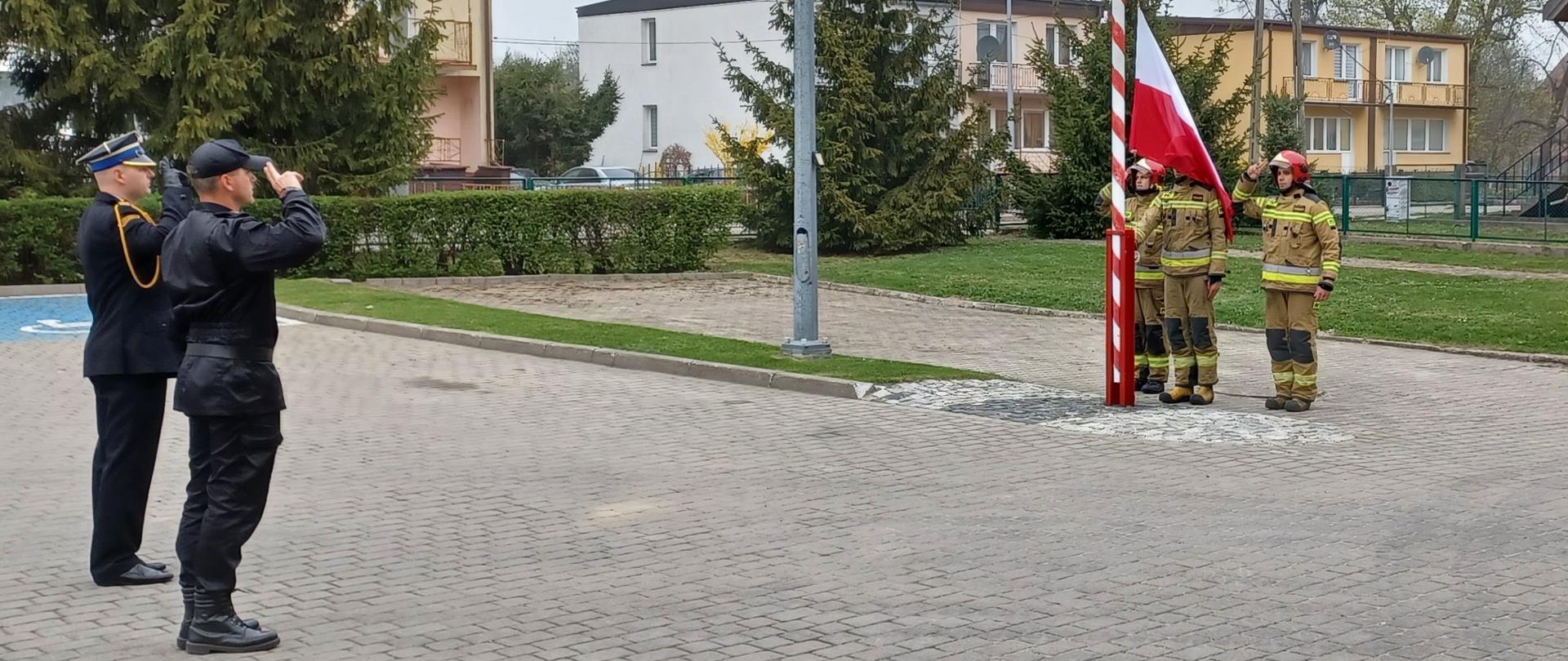 Na placu przed KP poczet flagowy podnosi flagę na maszt. Na przeciwko flagi stoi Komendant wraz z dowódcą uroczystości i oddają honor. W oddali widać budynki mieszkalne. 