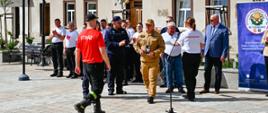 Komendant Miejski ubrany w piaskowy mundur wręcza statuetkę uczestnikowi zawodów ubranemu w czerwony podkoszulek z napisem straz i czarne spodnie. Z tyłu stoi kilkanaście osób w tle budynki w zabudowie szeregowej