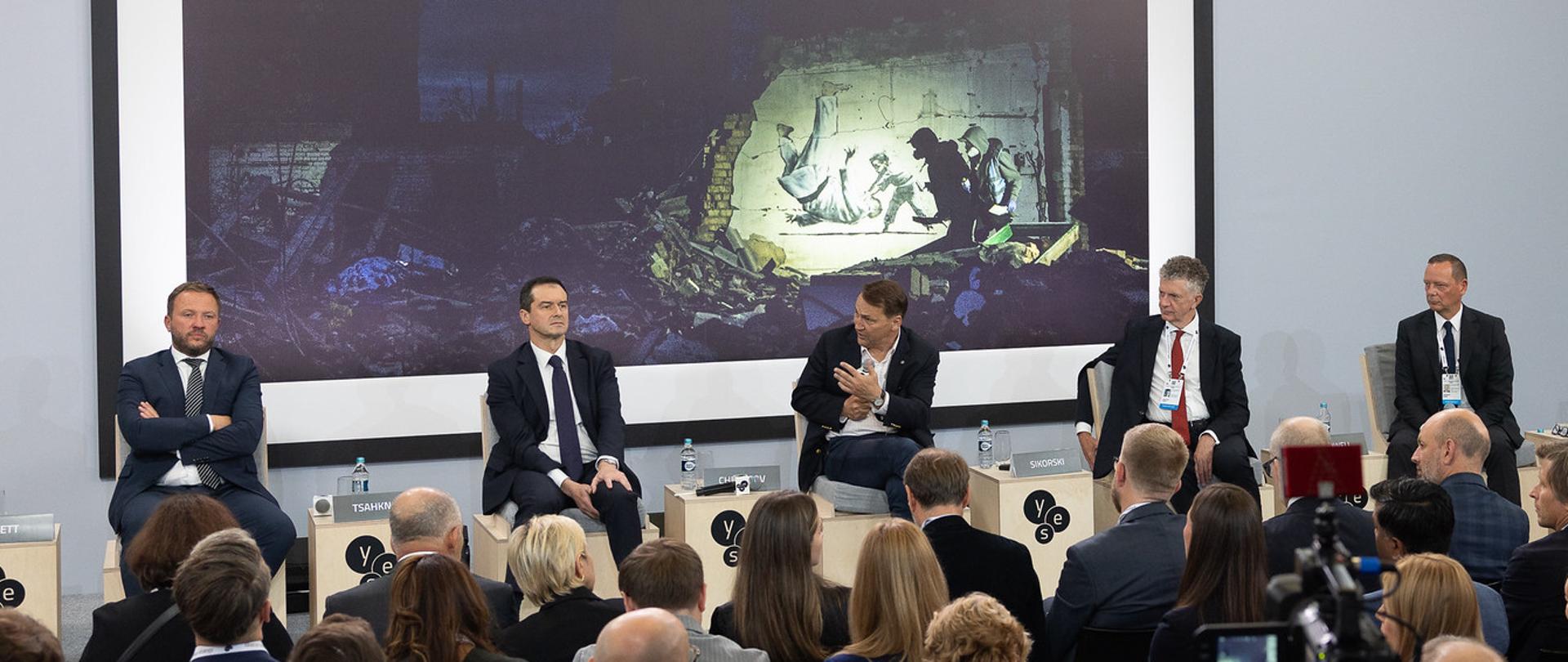 Deputy Prime Minister Radosław Sikorski takes part in a panel discussion with several men seated on stage in front of an audience. A large screen behind them displays an image of war damage, while the words “PEACE THROUGH STRENGTH NOW” appear above the stage; on the right, a live image of one of the panelists is displayed.
