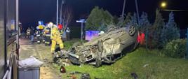Widać uszkodzony samochód po dachowaniu, który jest w rowie, jest noc, w oddali jest strażak