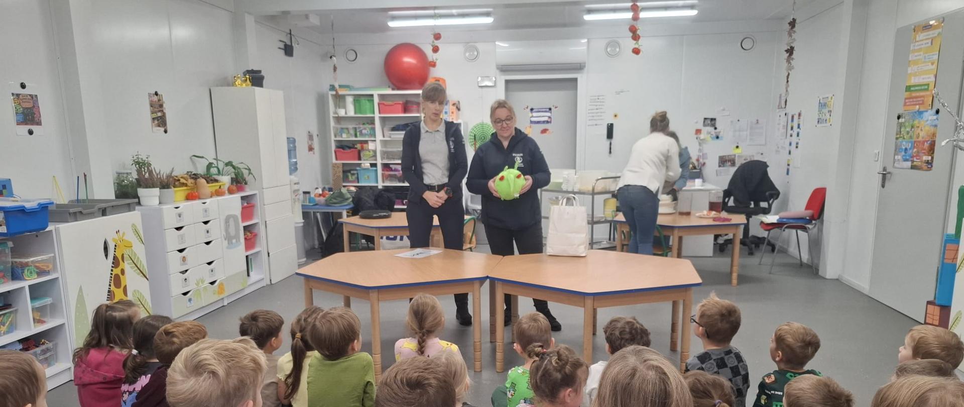Dwie kobiety stoją przed grupą przedszkolaków siedzących na podłodze w sali przedszkolnej. Jedna z kobiet trzyma zieloną maskotkę w kształcie szkodnika. W tle znajdują się stoliki i pomoce dydaktyczne.