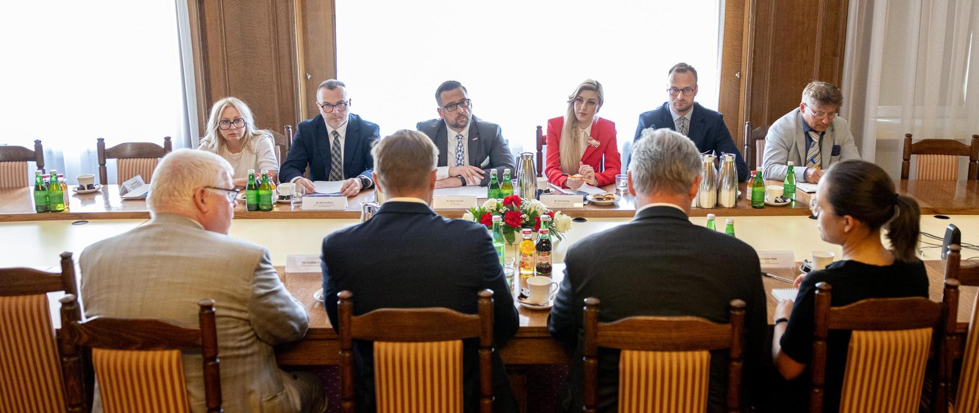 Przy stole siedzi grupa osób. Na stole stoją kwiaty, filiżanki i butelki. Widok na okno. 