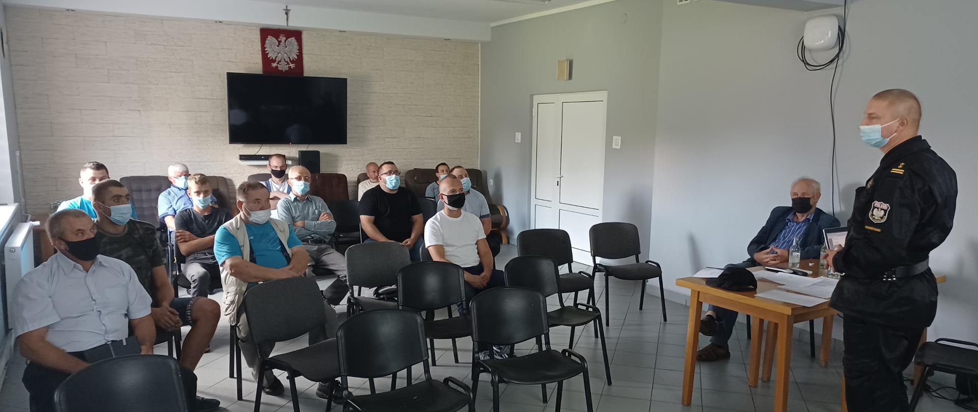 Zdjęcie obrazuje pomieszczenie, w którym ustawione są w pięciu rzędach krzesła w kolorze czarnym. Na krzesłach siedzą mężczyźni jest ich łącznie zgromadzonych 15 osób jest również kilka krzeseł wolnych w pierwszym rzędzie. Osoby posiadają zakryte usta i nos maseczkami. Po prawej stronie zdjęcia frontem do siedzących mężczyzn stoi kolejny mężczyzna ubrany w umundurowanie służbowe, dwuczęściowe koloru czarnego z przepiętym czarnym pasem skórzanym. Mężczyzna stoi obok drewnianego stołu, na którym stoi laptop oraz są poukładane dokumenty. Pomieszczenie posiada jedną ścianę pomalowaną na kolor szary w tej ścianie zamontowane są również białe drzwi, natomiast druga ściana za siedzącymi mężczyznami jest wyłożona w dekoracyjne cegiełki koloru kremowego. Podłogi wyłożone są terakotą koloru jasno niebieskiego.